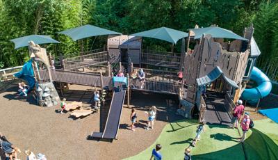 A large nature themed play structure filled with playing families. Extra wide inclusive accessible ramps connect the raised playground decking to different play experiences. The fenced in play area butts up next to a lush green woods.