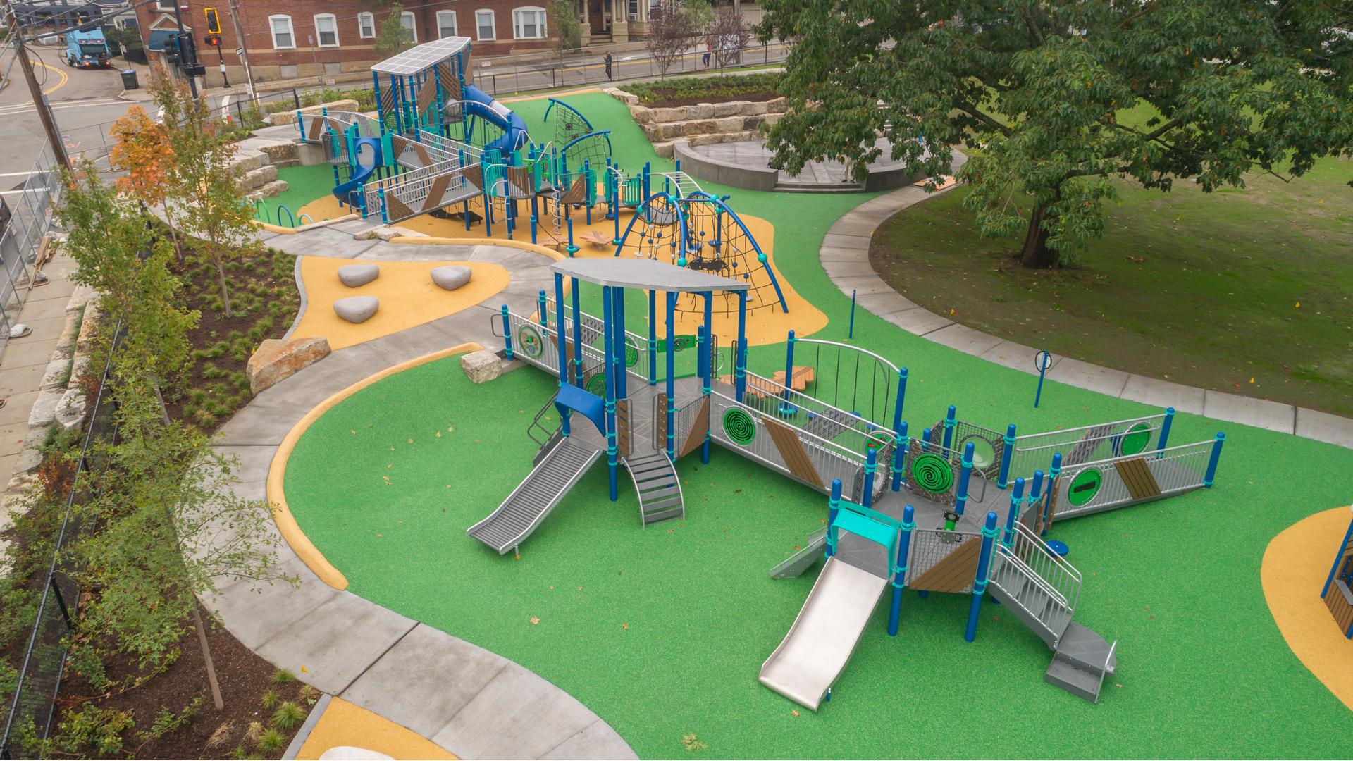 Elevated view of a outdoor play area with one play structure accessible by ramps and open playground decking, slides, climbers and play panels while the other has multiple unique rope climbers, ramps, slides, and play panels.