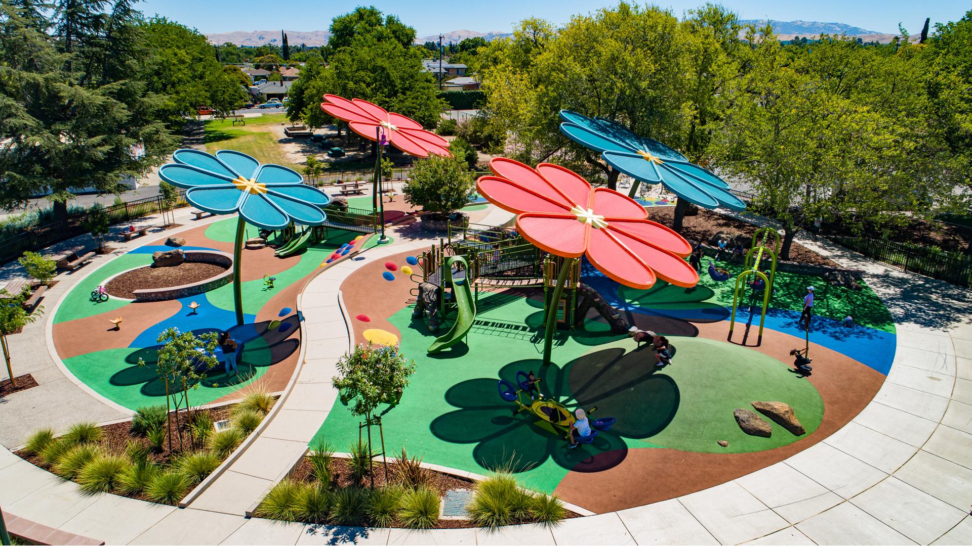 Four large red and blue shades designed like bloomed daisy flowers cover a large colorful play area with playground and other freestanding play activities for all abilities. 