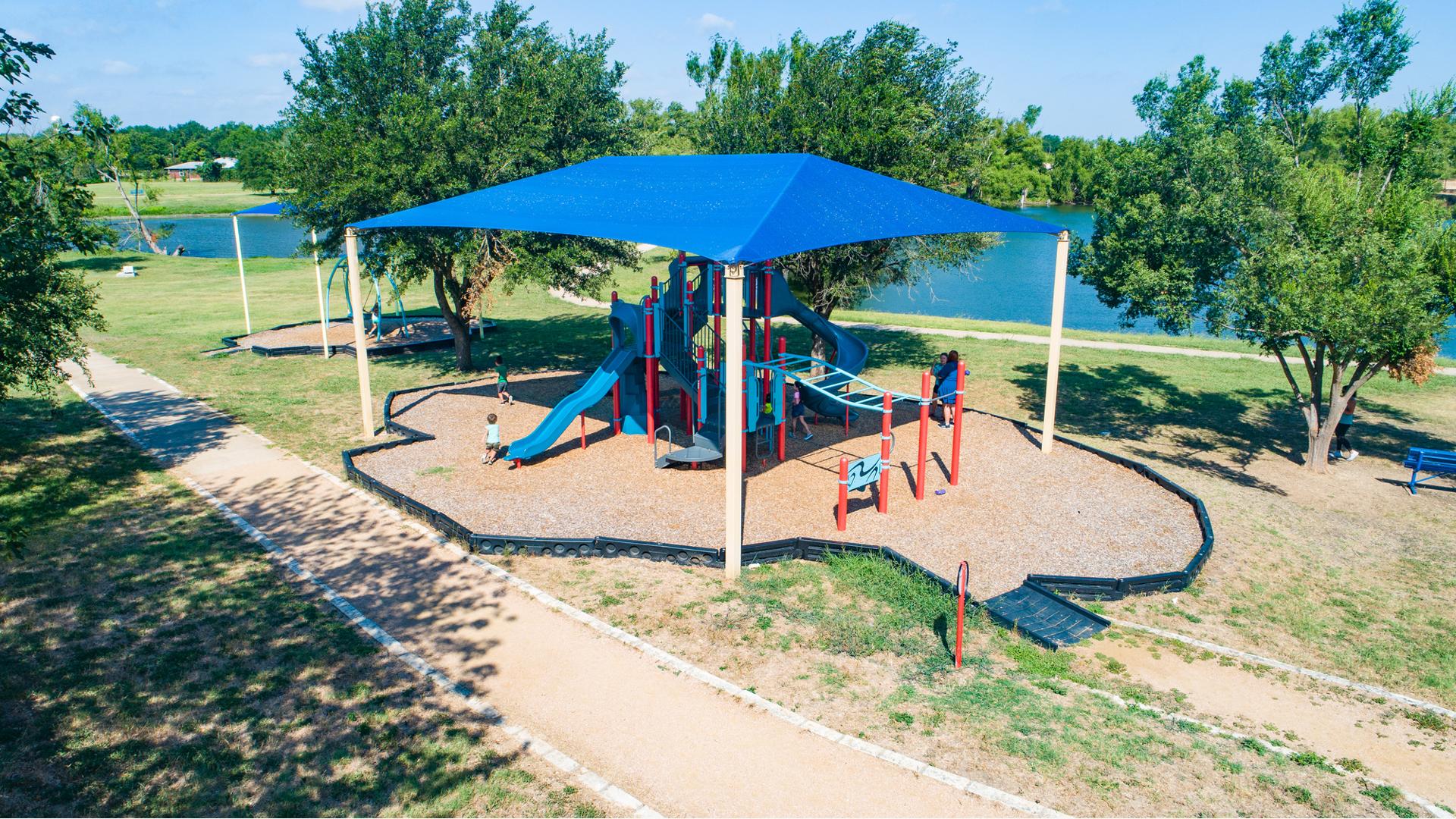 Elevated view of a large blue rectangular shade over a play structure all set next to a nearby lake.