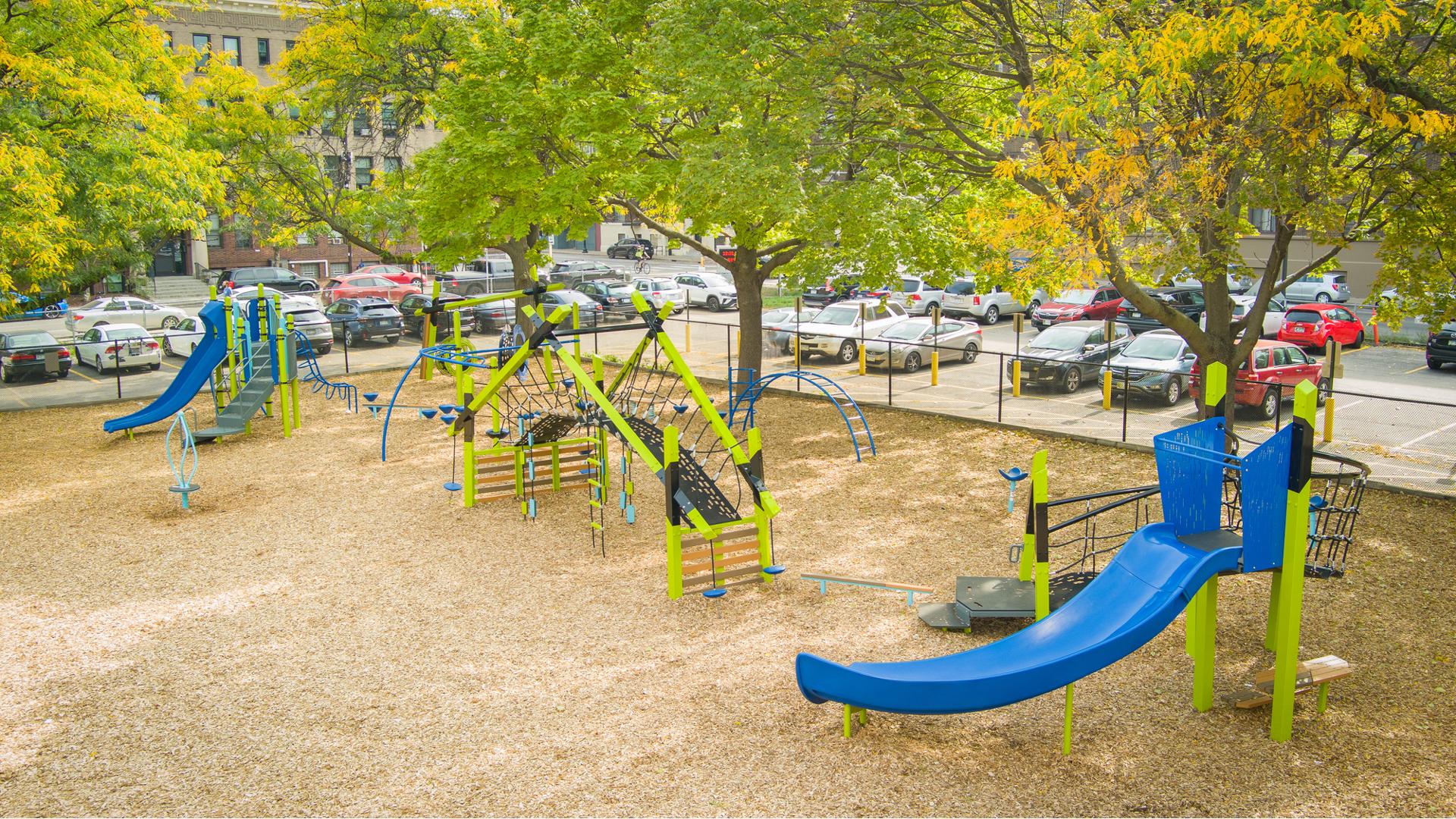 Elevated view of a city elementary school playground with three separate main play structures surrounded by mature trees and a parking lot.