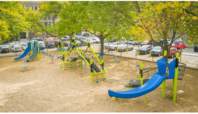 Elevated view of a city elementary school playground with three separate main play structures surrounded by mature trees and a parking lot.