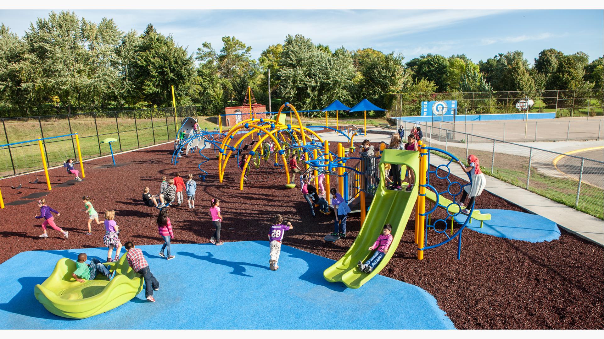 A class of children play on the brightly colored yellow arched posts and blue spinners and climbers of a large school playground surrounded by metal fencing.