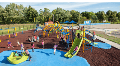 A class of children play on the brightly colored yellow arched posts and blue spinners and climbers of a large school playground surrounded by metal fencing.