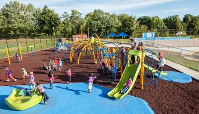 A class of children play on the brightly colored yellow arched posts and blue spinners and climbers of a large school playground surrounded by metal fencing.