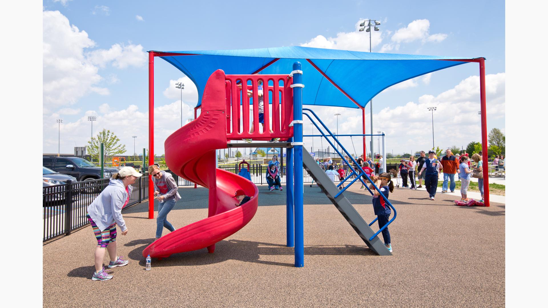 Two women watch as a boy slides down a spiral freestanding playground slide with a shade structure in the background