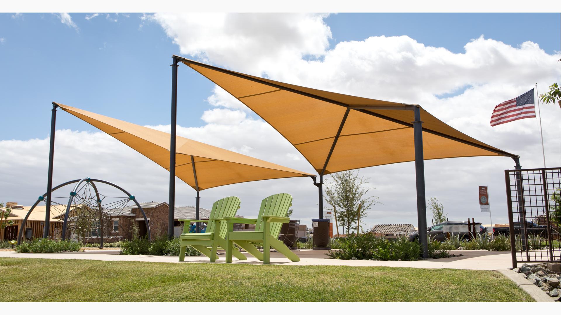 A pair of green lawn chair sit in front of a playground climber shaded by two peak side-by-side tan shade structures.