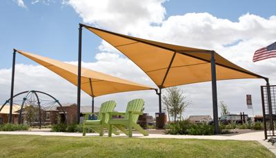 A pair of green lawn chair sit in front of a playground climber shaded by two peak side-by-side tan shade structures.