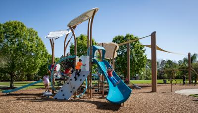 Children play on an air themed play structure at a park playground. Children climb on different climbers as a girl rides down the large blue slide of the structure.