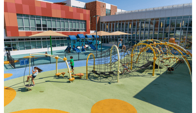 A large play area set in the inlet of a large building. A black iron fence separates a play area for younger children and older children. In the foreground older children play on a custom obstacle course like rope maze connected by large yellow arched posts.