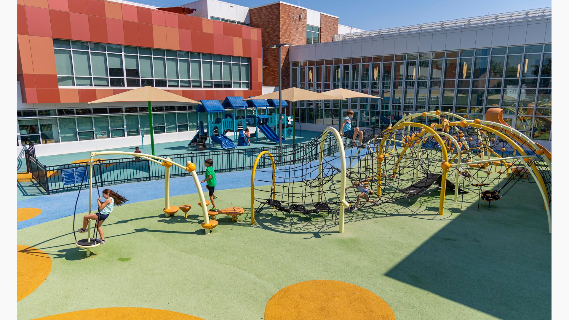 A large play area set in the inlet of a large building. A black iron fence separates a play area for younger children and older children. In the foreground older children play on a custom obstacle course like rope maze connected by large yellow arched posts.