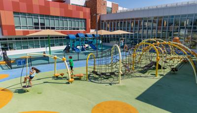 A large play area set in the inlet of a large building. A black iron fence separates a play area for younger children and older children. In the foreground older children play on a custom obstacle course like rope maze connected by large yellow arched posts.