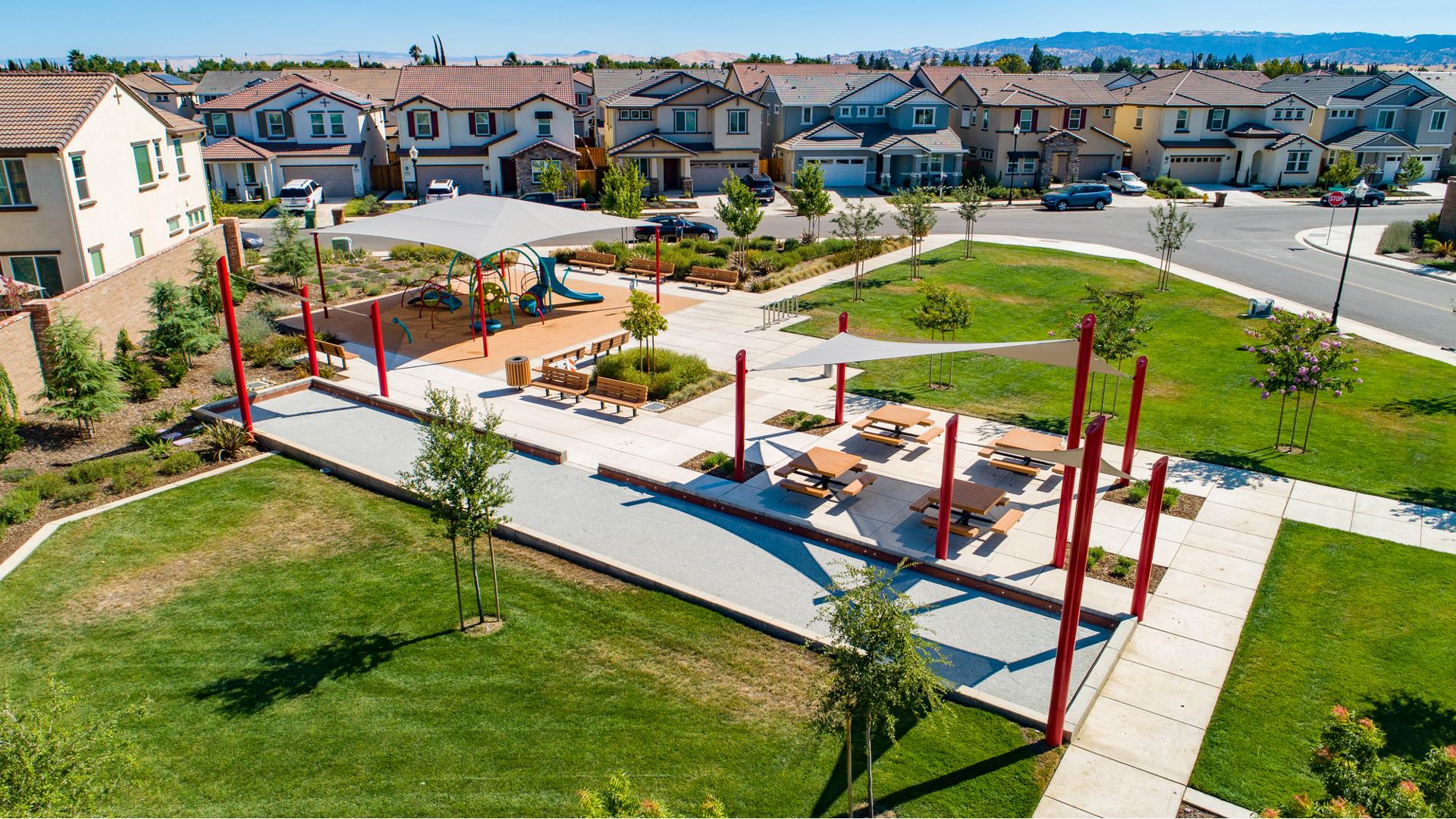 Elevated view of a neighborhood park with square play area with large shade connected by concrete pathways to a seating area with picnic tables next to a bocce ball court. 