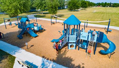 Park playground nestled near open grassy soccer fields features a zipline, 3 bays of swings, and a bright blue traditional playground for toddlers and a complementary school-age playground filled with twisty slide, interactive panels and tons of climbers.