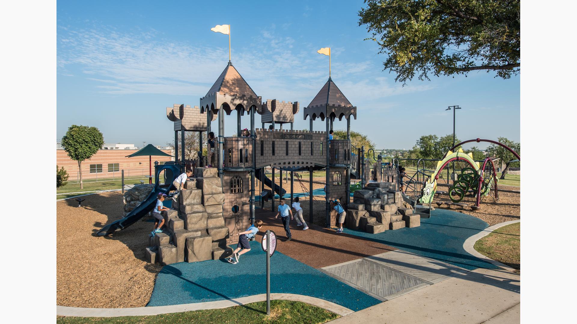School aged children wearing school uniforms playing on castle-themed playground. Play space also includes a dragon themed playground. 