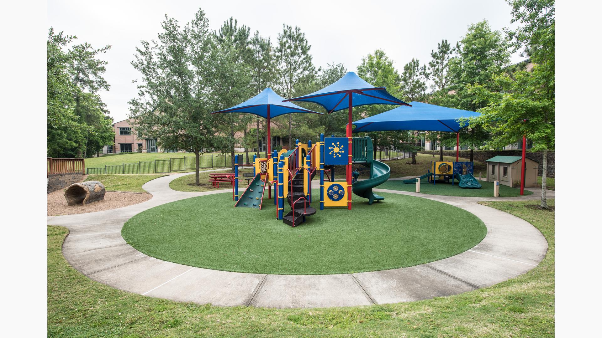 View from the top of a small hill down to a play area with a play structure with shades for older children with a smaller play structure to the right with a shade for toddlers all surrounded by lush green mature trees.
