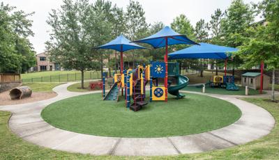 View from the top of a small hill down to a play area with a play structure with shades for older children with a smaller play structure to the right with a shade for toddlers all surrounded by lush green mature trees.