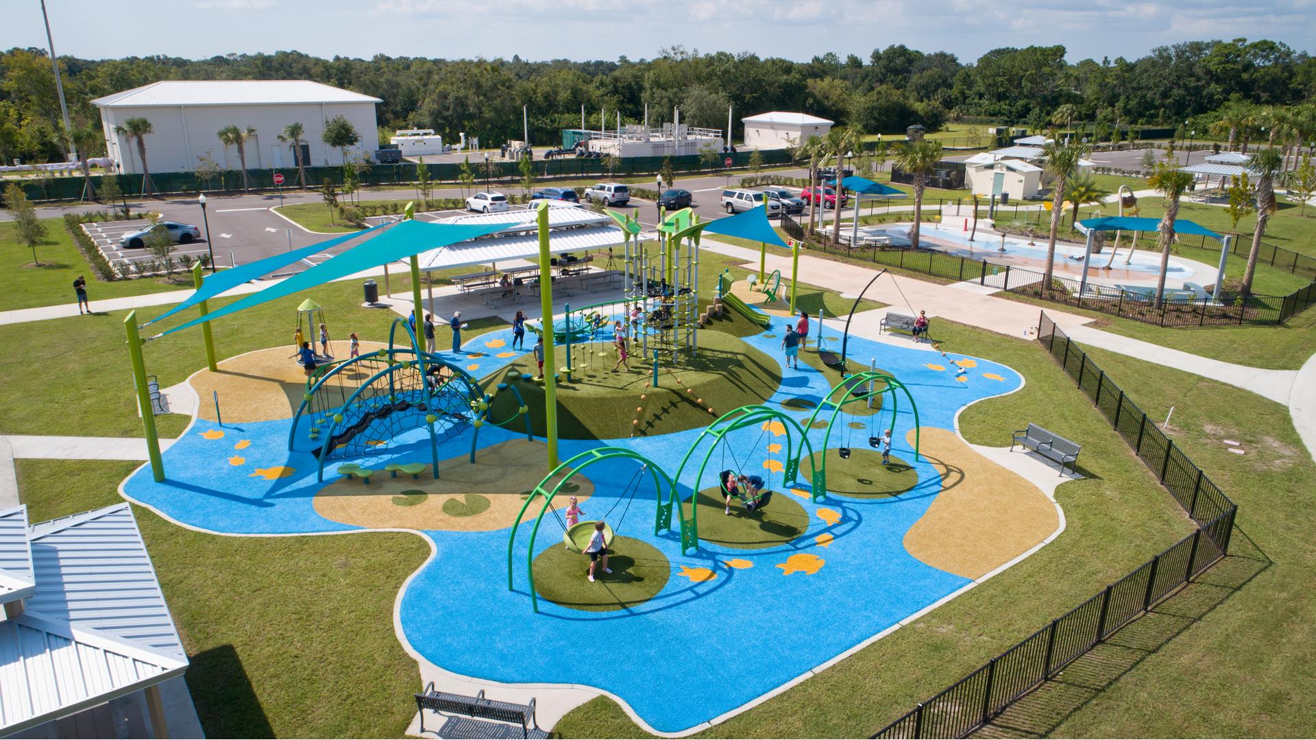 A playground filled with children designed with a pond theme showcasing lilly pads, fish, and nature all next to a nature themed splash pad. 