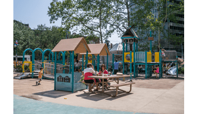 A mother on a bench has lunch with her son while visiting the Pearl Street Playground in New York City. Other parents watch as their children play on the playground in the background.