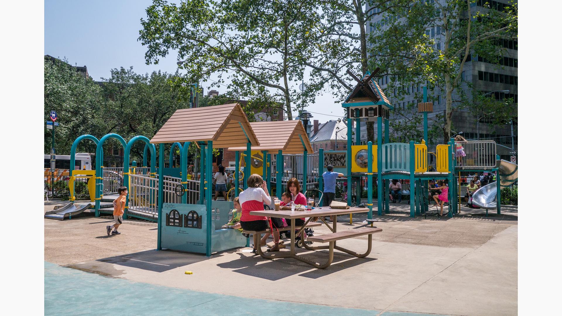 A mother on a bench has lunch with her son while visiting the Pearl Street Playground in New York City. Other parents watch as their children play on the playground in the background.