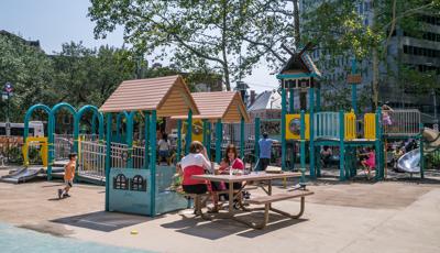 A mother on a bench has lunch with her son while visiting the Pearl Street Playground in New York City. Other parents watch as their children play on the playground in the background.