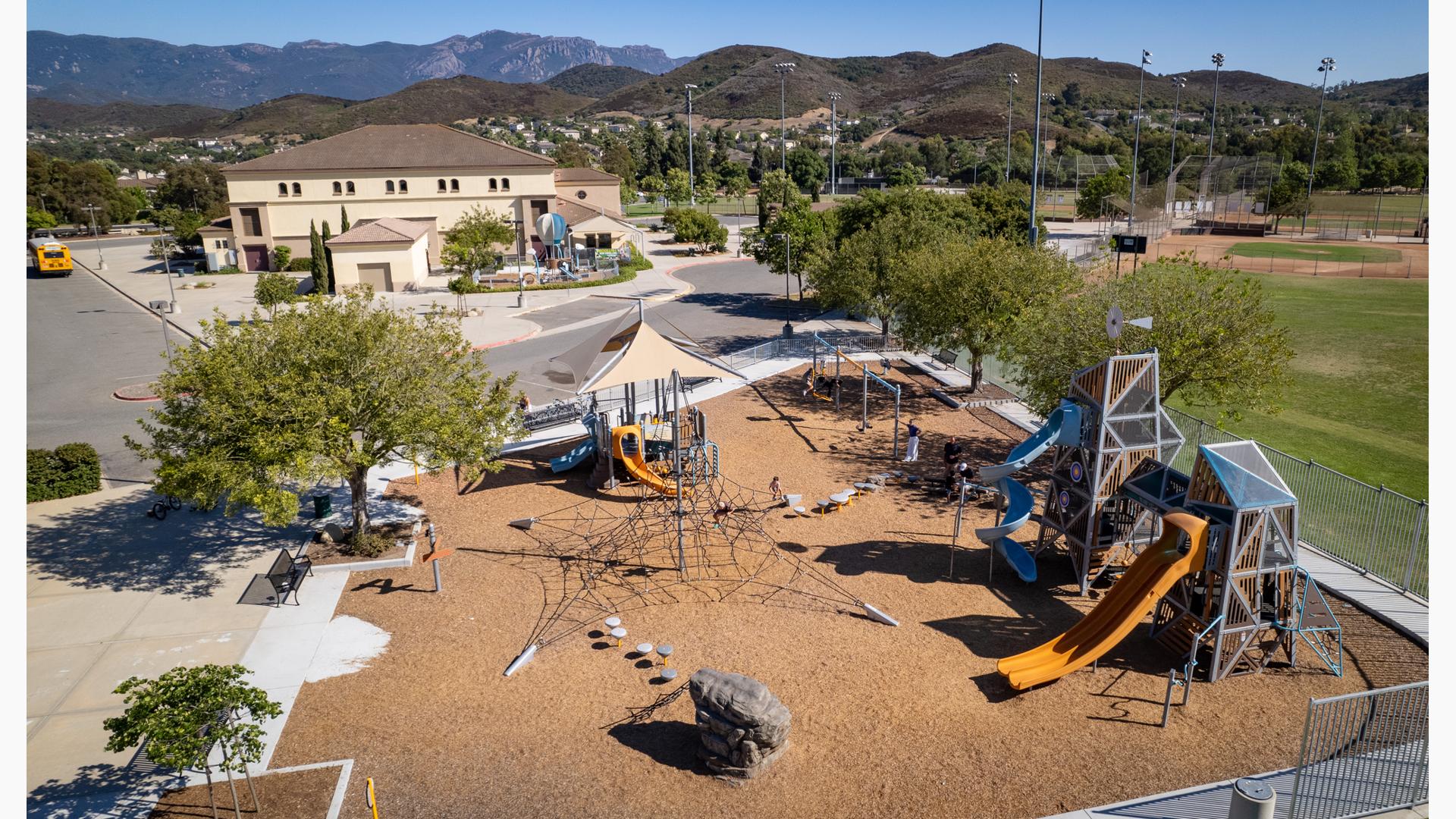 Elevated view of a park playground and an additional play structure across the street in a enclosed area for younger children.