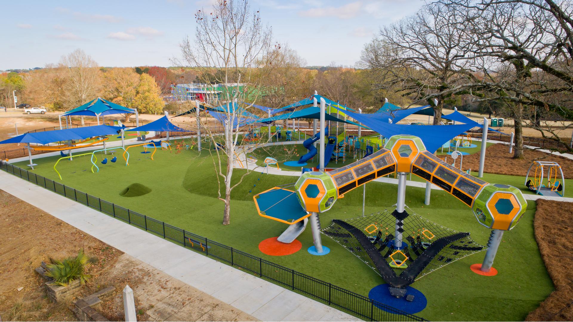 Elevated view of a play area surrounded by a black iron fence. Three towers with hexagonal play pods are connected by enclosed crawl tunnels in the foreground while triangular blue shade sails cover a large play structure and other play activities in the background.  