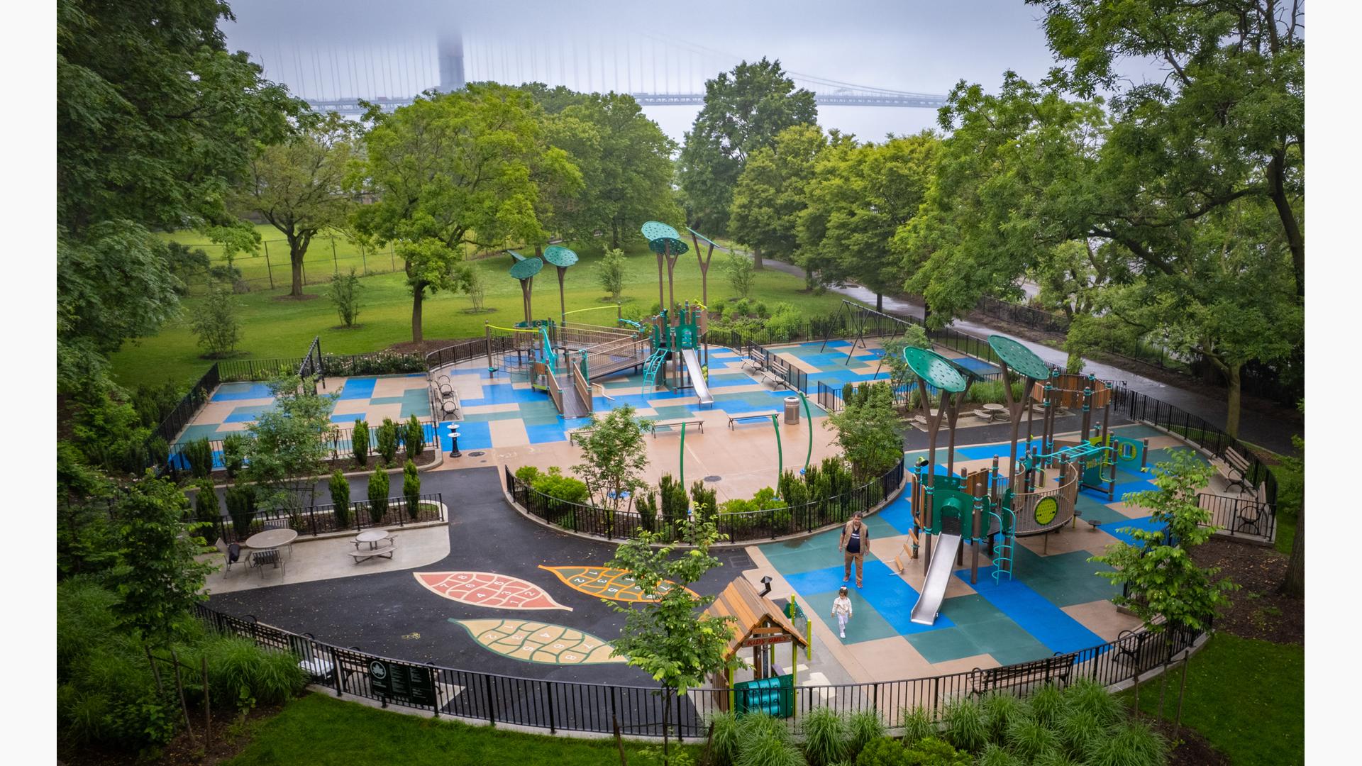 Elevated view of a park playground surrounded by large trees and a river front nearby.