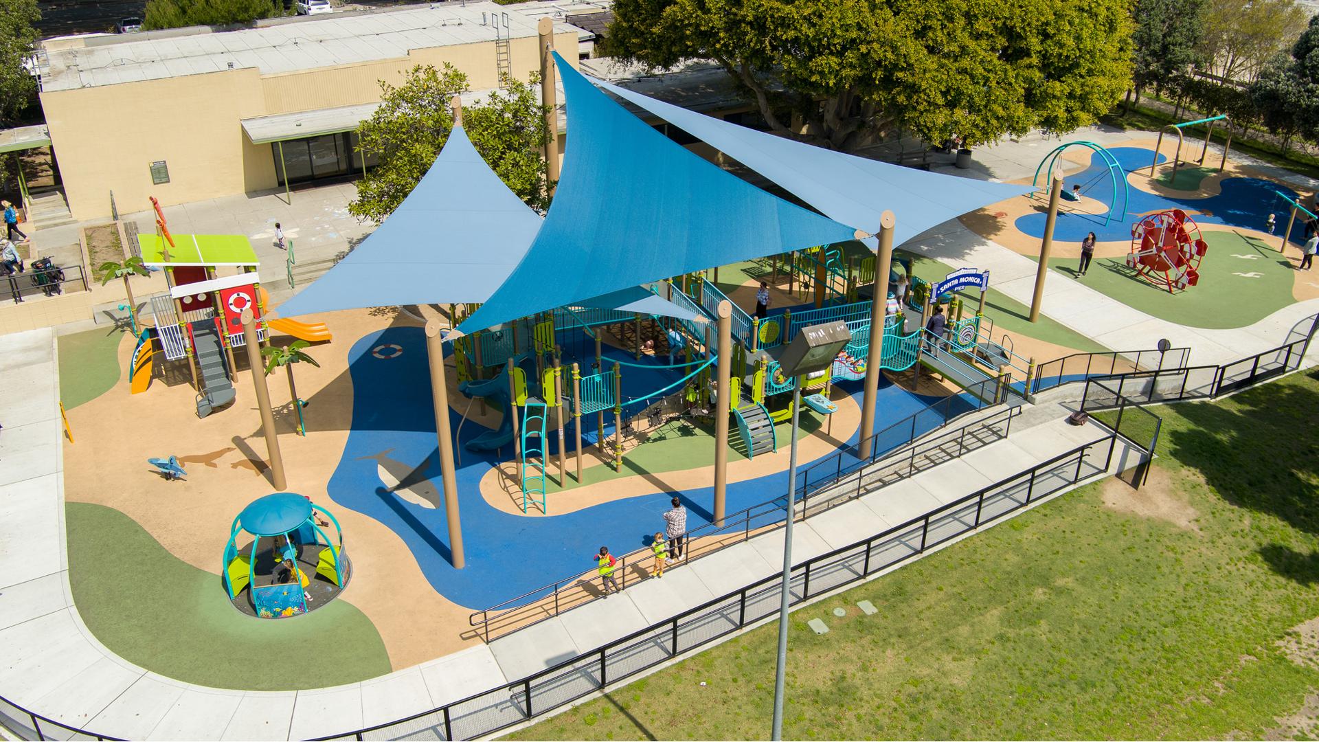 Elevated view of large blue triangle shade sails over a large inclusive beach themed playground. 