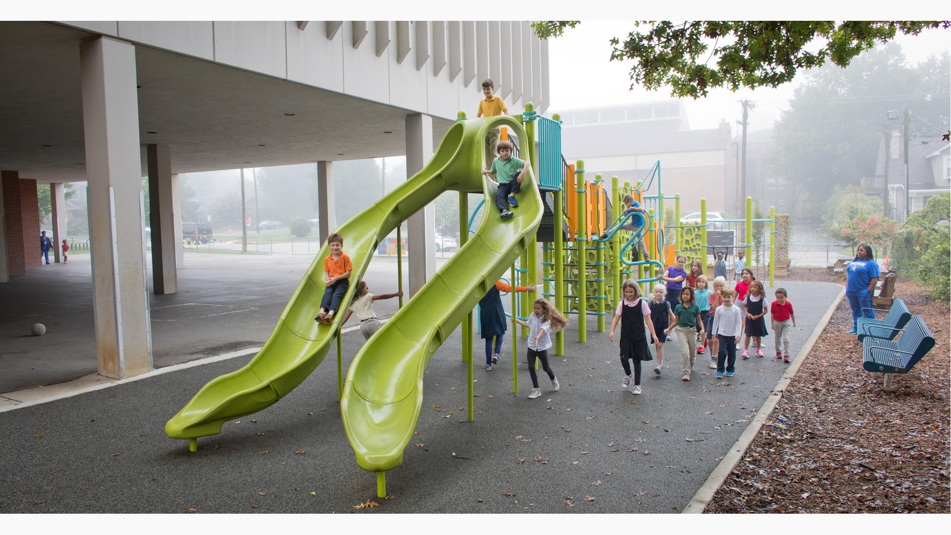 School kids playing in the misting rain on a playground with two limon colored slides in the foreground.