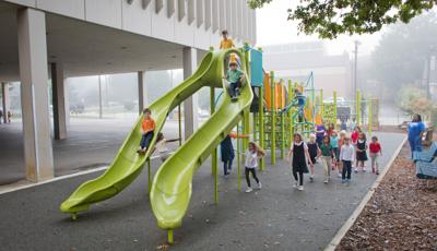 School kids playing in the misting rain on a playground with two limon colored slides in the foreground.