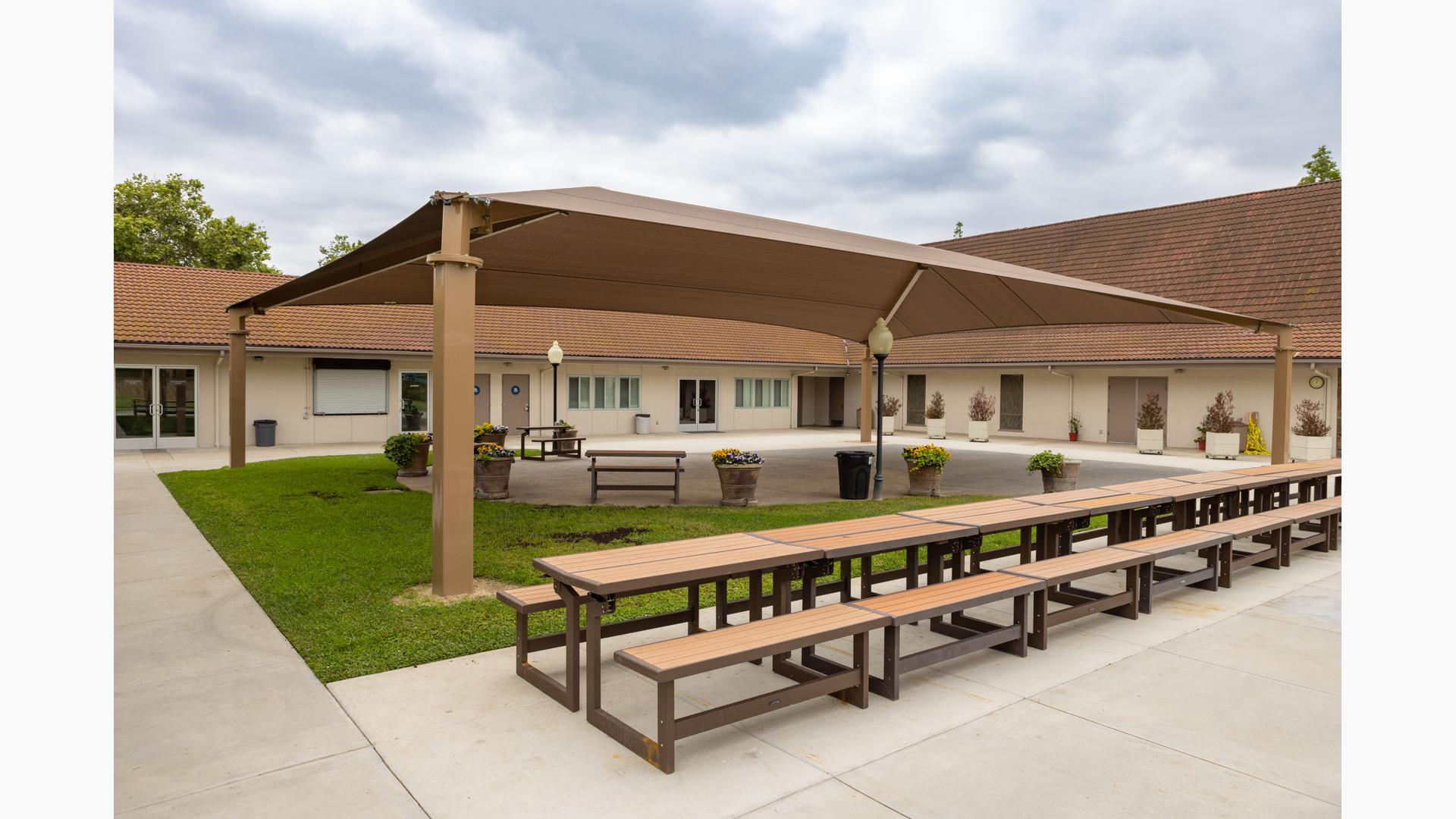 A center court yard with large brown rectangular shade pavilion with a row of picnic tables on one side.