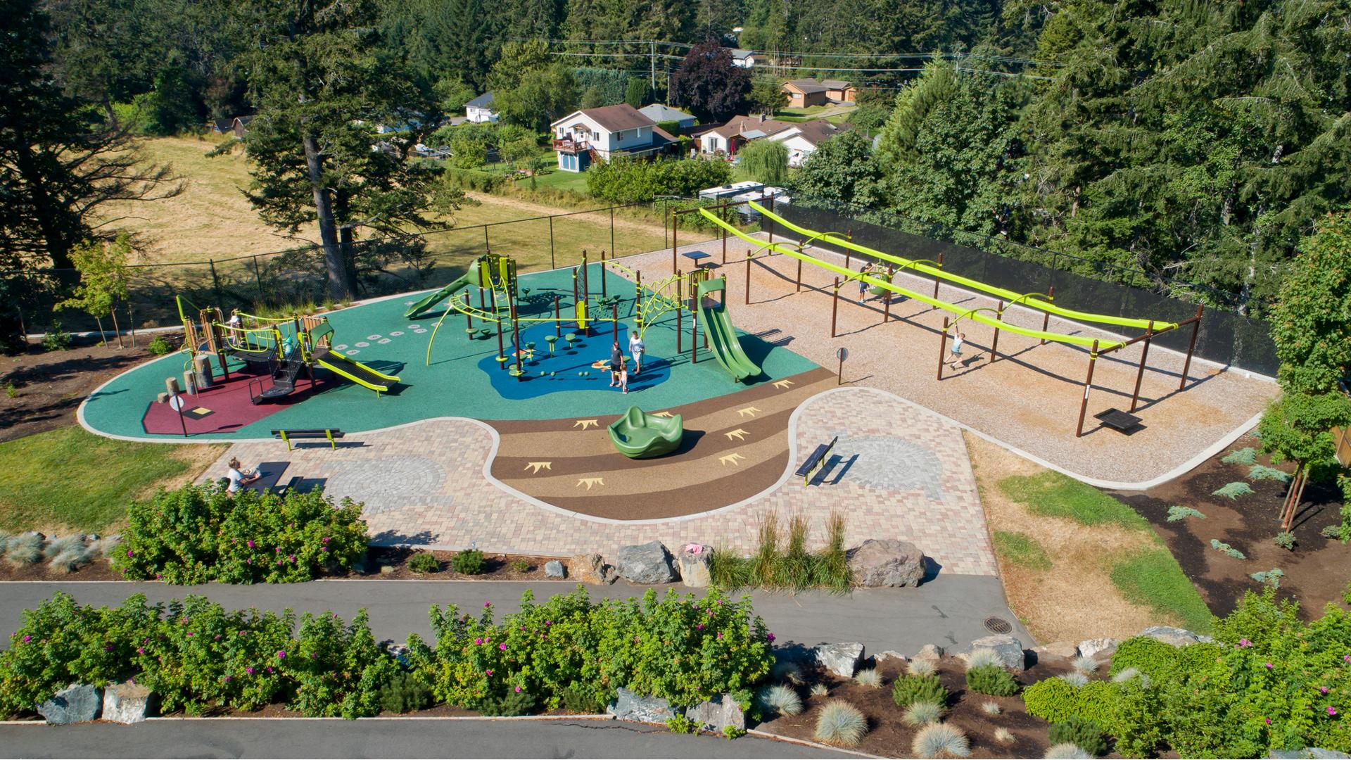 Full elevated view of a large park space. Children play on two separate play structures with many play activities while others play on two zip line bays.