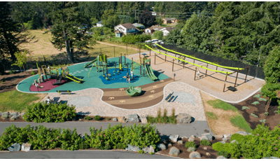 Full elevated view of a large park space. Children play on two separate play structures with many play activities while others play on two zip line bays.