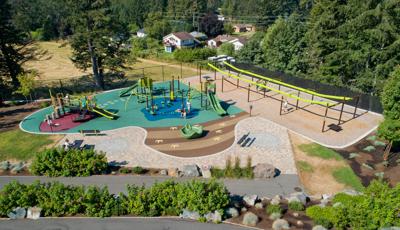 Full elevated view of a large park space. Children play on two separate play structures with many play activities while others play on two zip line bays.