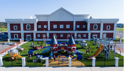 School children wearing school uniforms play on castle-themed playground structure. Childcare-aged children play on three playhouse structures colored red, tangerine and blue. 