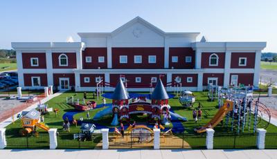 School children wearing school uniforms play on castle-themed playground structure. Childcare-aged children play on three playhouse structures colored red, tangerine and blue. 