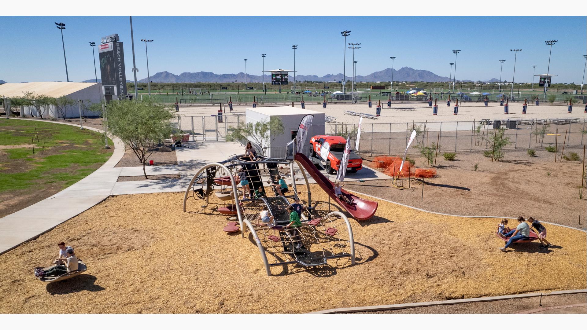 Elevated view of children playing on a uniquely wave shaped play structure made of ropes and belted pathways slides and climbers. Other children play on two separate play activities near the larger play structure. The background is filled with sand volleyball courts and baseball fields beyond that.