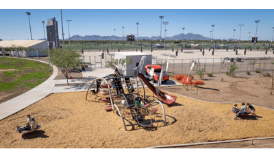 Elevated view of children playing on a uniquely wave shaped play structure made of ropes and belted pathways slides and climbers. Other children play on two separate play activities near the larger play structure. The background is filled with sand volleyball courts and baseball fields beyond that.