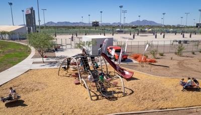 Elevated view of children playing on a uniquely wave shaped play structure made of ropes and belted pathways slides and climbers. Other children play on two separate play activities near the larger play structure. The background is filled with sand volleyball courts and baseball fields beyond that.