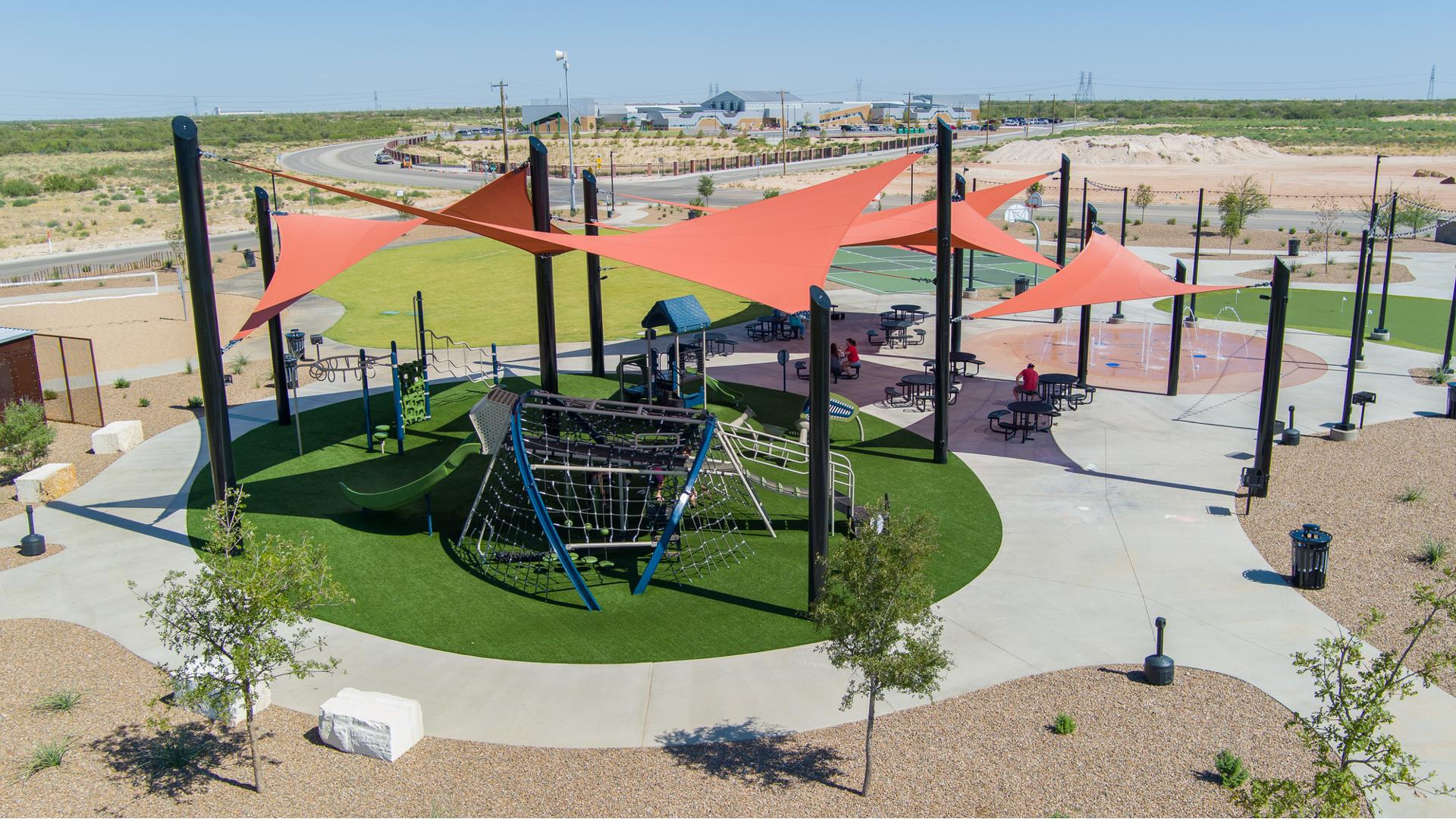 Elevated view of large orange triangular shade sails covering a circular play area with artificial grass and three separate structures to play on.