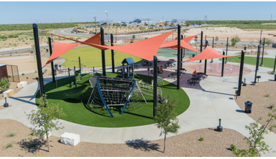Elevated view of large orange triangular shade sails covering a circular play area with artificial grass and three separate structures to play on.