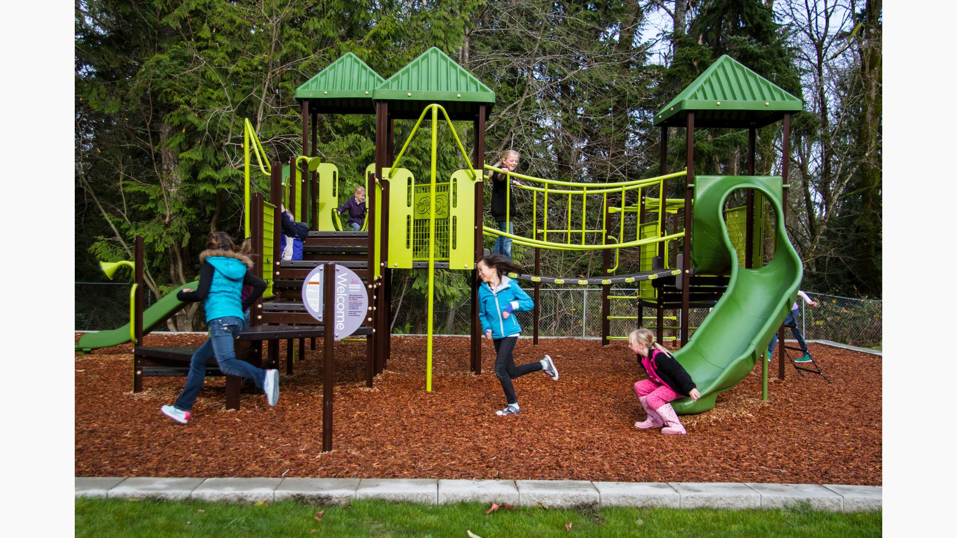 Kids playing on nature inspired playground with green roofs and play equipment. Brown stairs with linking bridge. Tall evergreen trees surround the playground while three girls play chase across the wood chips around the playground.