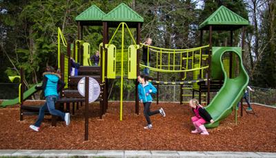 Kids playing on nature inspired playground with green roofs and play equipment. Brown stairs with linking bridge. Tall evergreen trees surround the playground while three girls play chase across the wood chips around the playground.