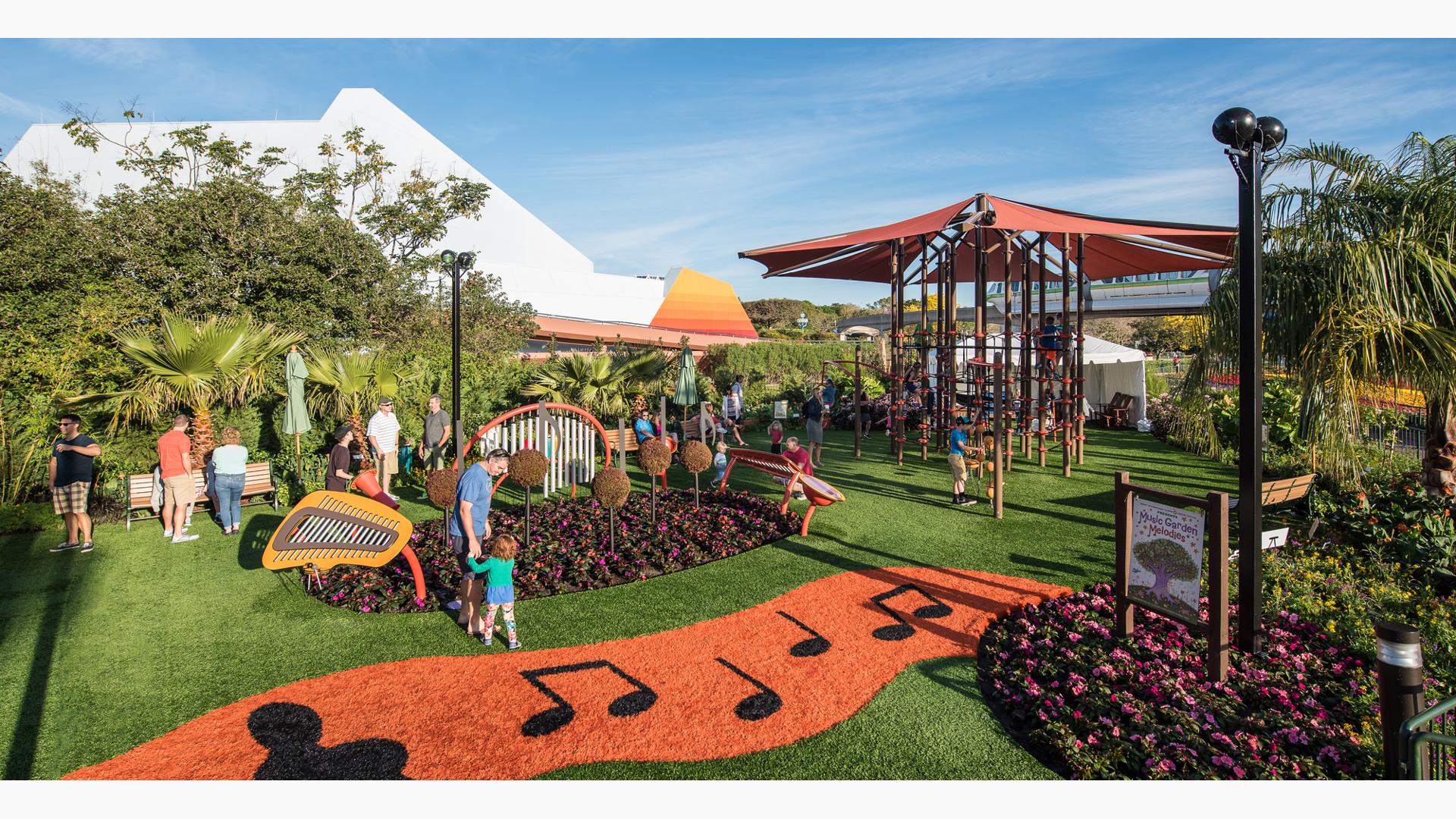 Families play on outdoor musical instruments scattered amongst a tropical garden with an additional large climbing tower structure with ropes and overhead shade.