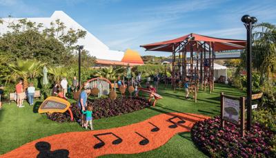 Families play on outdoor musical instruments scattered amongst a tropical garden with an additional large climbing tower structure with ropes and overhead shade.