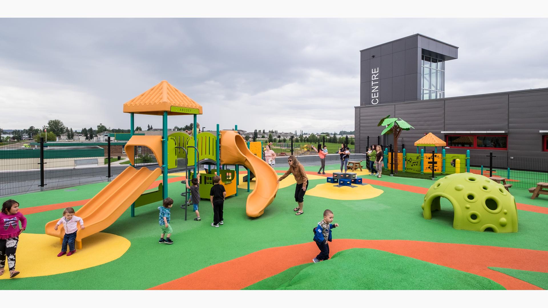 Children play at a fenced in play area next to a building. Two separate play areas are separated by a fence one area for older children and the other for younger kids. 