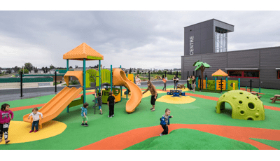 Children play at a fenced in play area next to a building. Two separate play areas are separated by a fence one area for older children and the other for younger kids. 