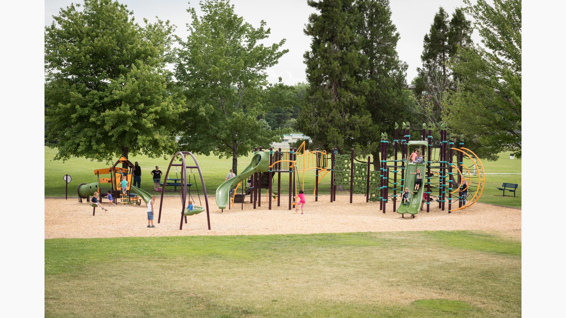 Families play at a park playground with play structures that have unique rope disc and geometric climbers, younger age appropriate play structures and a inclusive large disc swing.