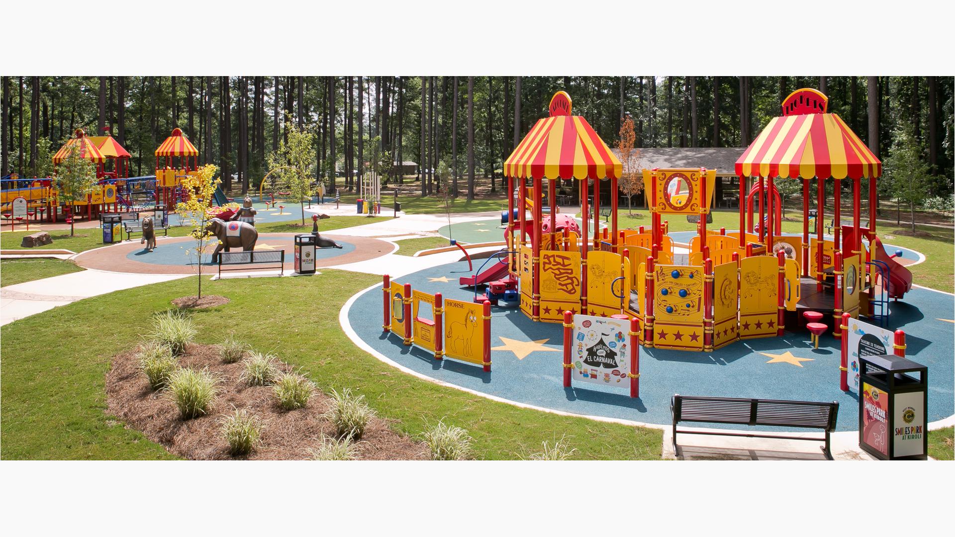In the foreground yellow and red colored circus themed play structure has roof panels designed like circus tents. A second circus themed play structure sits in the background along with a surrounding wall of tall trees.